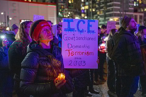 Protester holding a candle and a sign reading “I.C.E. Govt sanctioned domestic terrorism since 2003” during a nighttime vigil in Seattle, surrounded by a crowd with city lights in the background.