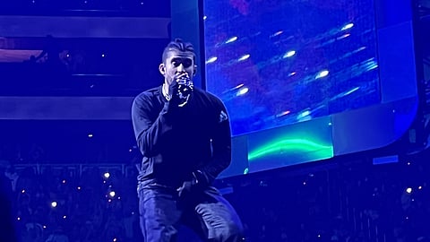 Musician performs on stage at Seattle’s Climate Pledge Arena, singing into a microphone under blue lighting with a large video screen behind him during a live concert in 2022.