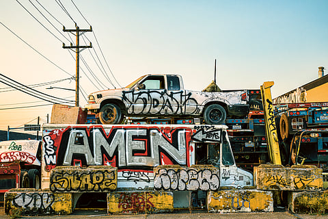 A photo shows a white pickup truck on top of a white delivery van, both vehicles, as well as surrounding structures, tagged with graffiti.