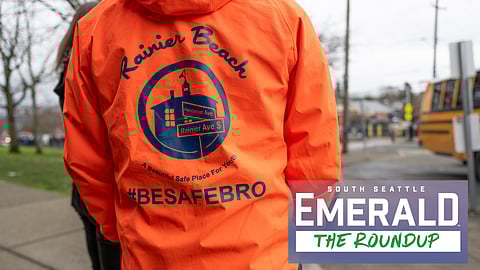 A person wears a bright-orange jacket. The photo shows the back of the jacket, which reads "Rainier Beach: A Beautiful Safe Place for Youth" and the hashtag "Be Safe Bro." A school bus is in the background.