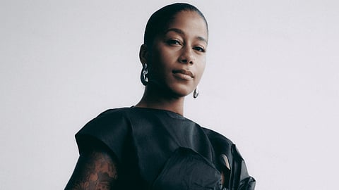 Poet and activist aja monet in a minimalist studio portrait, wearing a black structured top and hoop earrings, looking confidently at the camera against a light background.