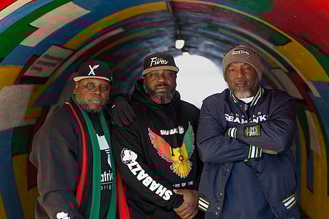 Three Black men standing shoulder to shoulder in a brightly painted tunnel, wearing caps, hoodies, and jackets.