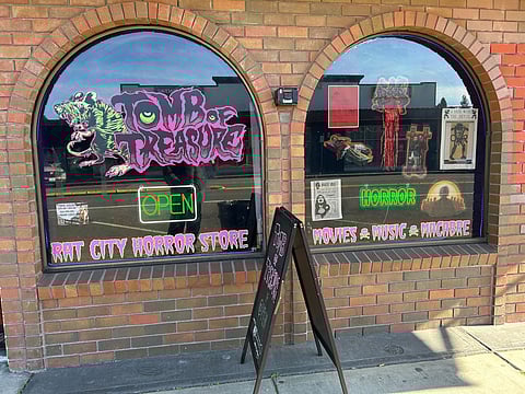 Rat City Horror Store storefront with arched brick windows, neon “Open” sign, and bold pink “Tomb of Treasure” graphic featuring a green rat, plus horror-themed posters and signage reading “Horror Movies Music Macabre.”