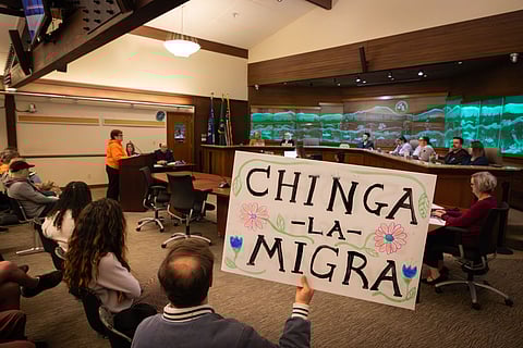 Tukwila City Council meeting with attendees seated as a speaker addresses officials; in the foreground, a person holds a handmade protest sign reading “Chinga La Migra” decorated with flowers.