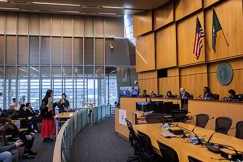 City council meeting in a large chamber as a speaker holds a “Shut Down ICE” protest sign while addressing officials seated at the dais, with audience members watching and U.S. and Washington state flags behind the council.