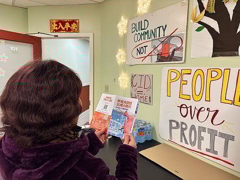Woman in a community center reads Chinese-language pamphlets beside posters saying “People Over Profit” and “Build Community Not Profit,” with water bottles and decorations on the wall.