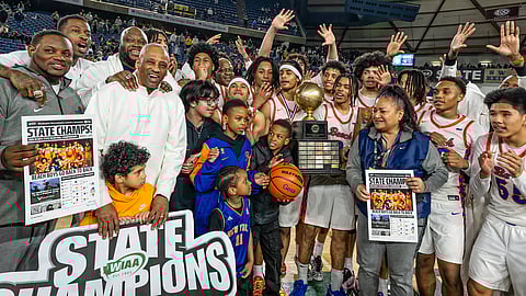 High school basketball team celebrates a WIAA state championship on the court, holding a trophy and “State Champs” signs while teammates, coaches, and families gather around.