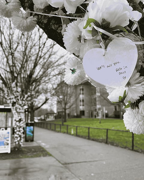 White flowers are strung around a tree next to a bus stop with a paper heart that reads, "We'll miss you. Rest in peace. <3"