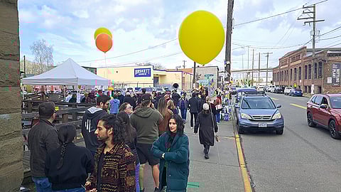 Crowd lined up along a city sidewalk at a street festival, with large yellow and orange balloons above tents and food stalls, cars passing on the road beside an industrial brick building.