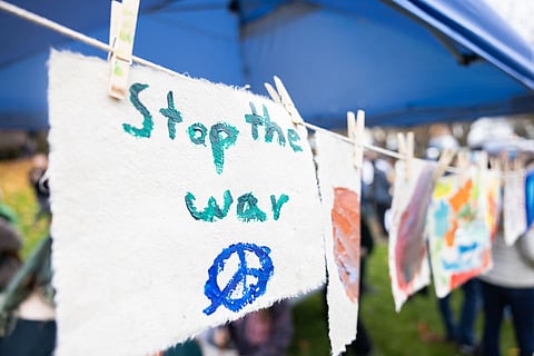 A handmade sign with the words "Stop the war" painted in teal alongside a blue peace symbol, hangs from a clothesline under a blue canopy, with a blurred background of protestors and children's artwork.