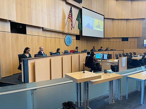 City council meeting with officials seated at a long dais, microphones and nameplates visible, U.S. and state flags behind them, and a large screen displaying a virtual participant.