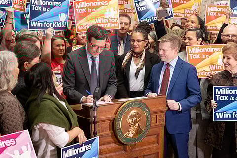 Politician signs “Millionaires Tax” bill at podium with Washington state seal, surrounded by supporters holding colorful “Win for Kids” and “Working Families” signs in a crowded indoor event.