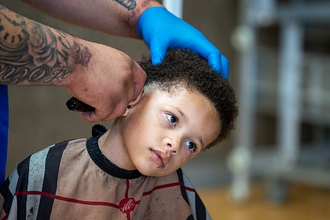 A young Black boy tilts his head as a barber uses clippers to trim the youth's hair.