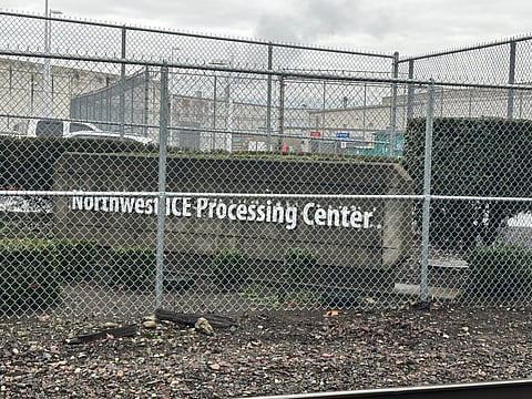 Chain-link fence in front of the Northwest ICE Processing Center sign, with detention facility buildings and overcast sky in the background.