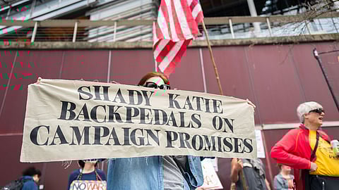 A protester holds a banner reading “Shady Katie backpedals on campaign promises” during a rally.