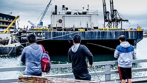 Anglers stand along a bridge railing, fishing beside cranes and a docked barge.