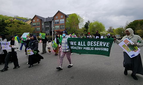 A group of people, two of which hold a banner proclaiming "We all deserve health public housing" march down a street.
