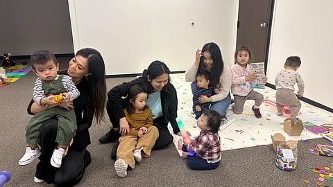 Group of women and young children sitting and playing together on a classroom floor with toys, books, and colorful mats.