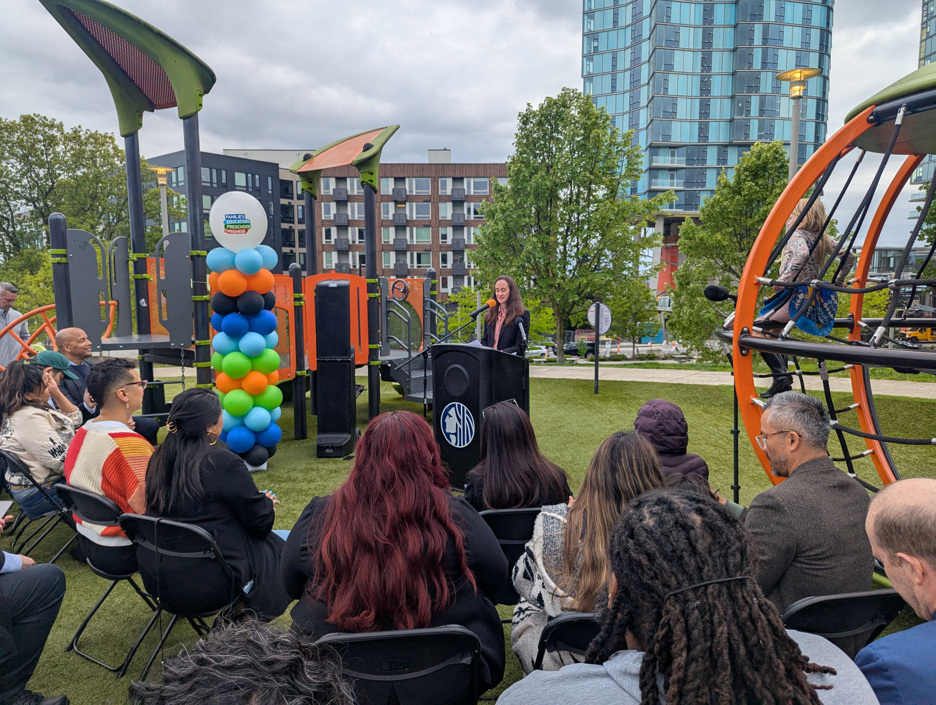 Woman speaking at a podium during an outdoor press event at a modern playground.