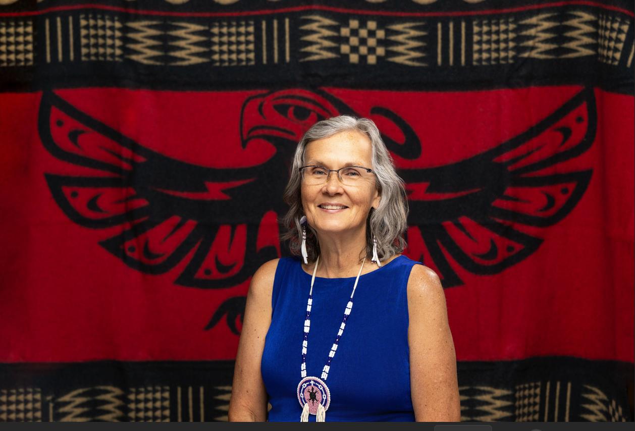 An Indigenous woman with gray shoulder-length hair, wearing a blue sleeveless dress and a beaded necklace, stands in front of a red blanket with an image of an eagle.