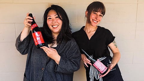 Two smiling women holding sake bottles against a light brick wall.