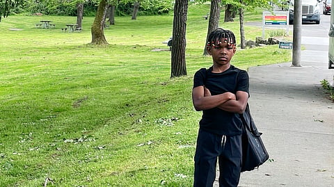A young Black youth with cornrowed hair and arms crossed stands near a grassy field.