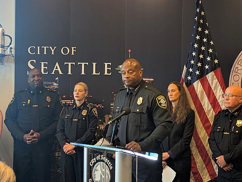 A Black man stands at a podium after being named interim police chief, and the mayor and other officers stand behind him near an American flag.