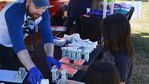Volunteer wearing blue hands juice boxes and prepared lunch to waiting youth.