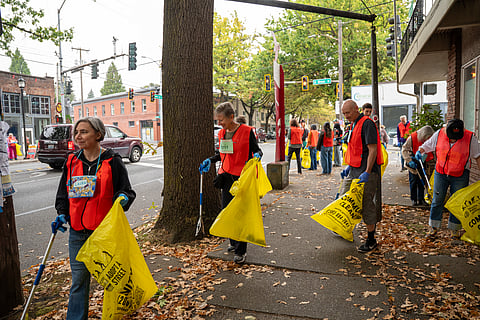 People in orange safety vests carry yellow trash bags as they clean up debris on the sidewalk.