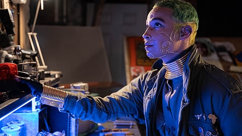 A person with dyed hair and space-age jewelry stands before a lit desk.
