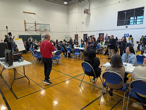 Eddie Lin in a red shirt speaks to people gathered in a community center gym.
