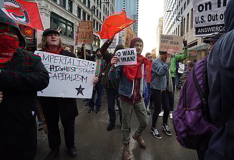 Anti-war protesters march through a city street holding signs reading “No War on Iran,” “Hands Off Iran,” and “Imperialism: Highest Stage Capitalism,” with red flags and banners raised.
