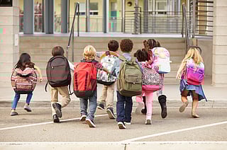 Children photographed from behind, all of them wearing backpacks and running toward a school.