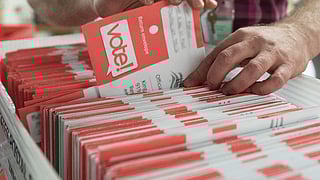 Close-up of a person sorting a large batch of official ballot envelopes labeled “vote!” in bold red letters. The envelopes are organized in a bin, part of a ballot processing operation, likely at an election office.
