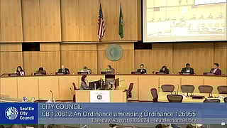 Seattle City Chambers with light natural wood walls and nine seated City Council members.  A person is typing at a desk in the foreground.