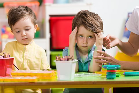 A grumpy looking kindergarten student sitting at a desk with art supplies with another student. 