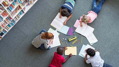 5 children around 6 years old lay and sit on a gray carpet next to a bookshelf. The children are coloring and drawing with colored pencils. 