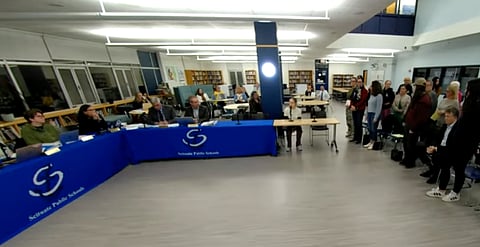 a meeting is being held in a library with the school committee seated on the left at a table and a group people stands up from chairs on the right 