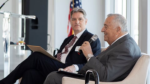 Two older men sit on a stage one has a microphone