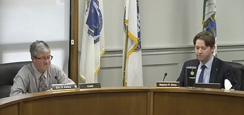 Two men at a desk with flags in the background