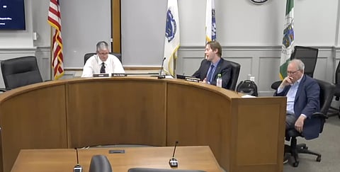 3 men sit at a round desk in a meeting room with flags behind them 
