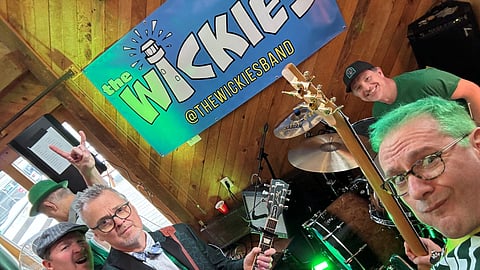 Members of Scituate band The Wickies pose in front of a band sign while holding their instruments.