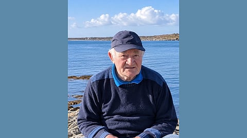 picture of older white man sitting on rocks by the coast wearing a navy blue sweater and navy blue baseball cap 
