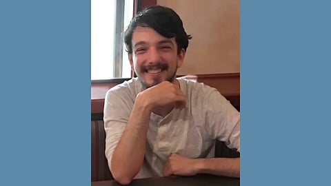 photo of smiling young white man with dark hair and a mustache and beard wearing a white button up shirt resting his chin on his hand