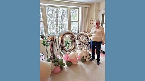 An white woman wearing jeans and a cream sweater stands smiling next to a large balloon display showcasing the number 108 