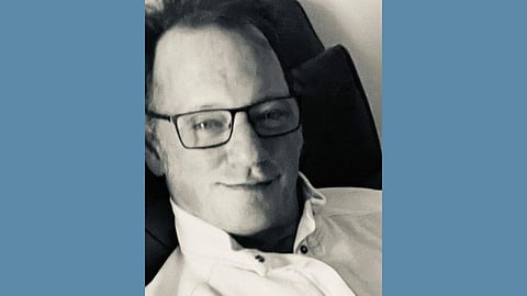 Black and white photo of a middle aged white man wearing black eyeglasses and a white button-up shirt with the collar standing up. He is smiling a closed mouth smile. 