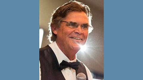 Photo of older white man with dark silver hair and eyeglasses, wearing a tuxedeo and holding a microphone, smiling warmly 