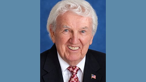 Photo of an elderly white man with white hair wearing a white shirt and red tie with a black blazer and an American flag lapel pin. He is smiling. 