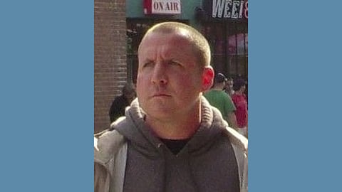 Photo of a middle aged white man wearing a grey hoodie and beige overcoat looking serious and staring into the distance 