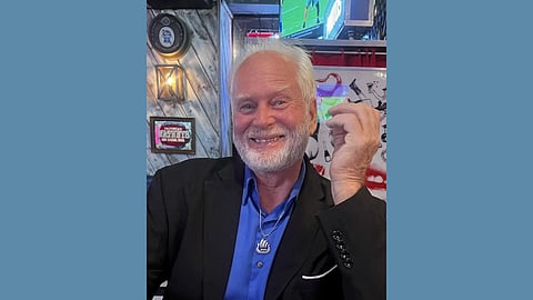 Photo of an older white man with white hair and beard wearing a blue button up shirt and black sport coat smiling warmly 